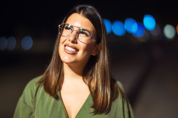 Young beautiful girl smiling happy and confident walking at the promenade, standing with a smile on face around night lights bokeh