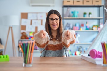 Young beautiful teacher woman wearing sweater and glasses sitting on desk at kindergarten pointing...
