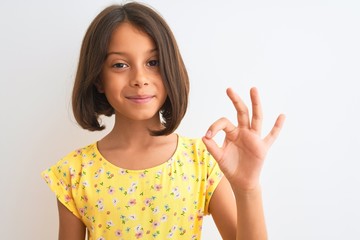 Young beautiful child girl wearing yellow floral dress standing over isolated white background doing ok sign with fingers, excellent symbol