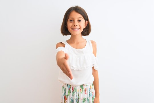 Young beautiful child girl wearing casual dress standing over isolated white background smiling friendly offering handshake as greeting and welcoming. Successful business.