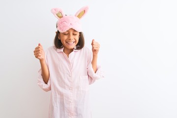 Beautiful child girl wearing sleep mask and pajama standing over isolated white background excited for success with arms raised and eyes closed celebrating victory smiling. Winner concept.