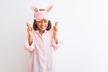 Beautiful child girl wearing sleep mask and pajama standing over isolated white background gesturing finger crossed smiling with hope and eyes closed. Luck and superstitious concept.