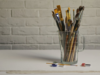 Brushes stand in a glass against a brick wall. On the table are watercolor paints