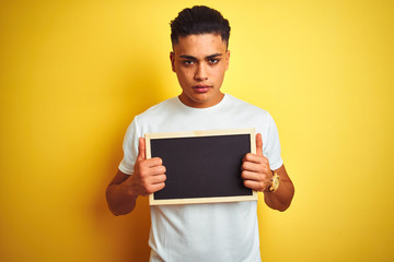 Young brazilian man holding blackboard standing over isolated yellow background with a confident expression on smart face thinking serious