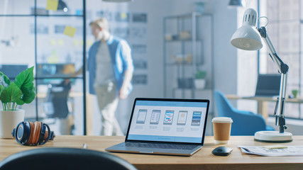 Shot of Laptop Standing at Desk in the Creative Office. Notebook Screen Shows Mobile Phone Application Design, Software UI Development. In Background Young Professional Stans in Creative Modern Office