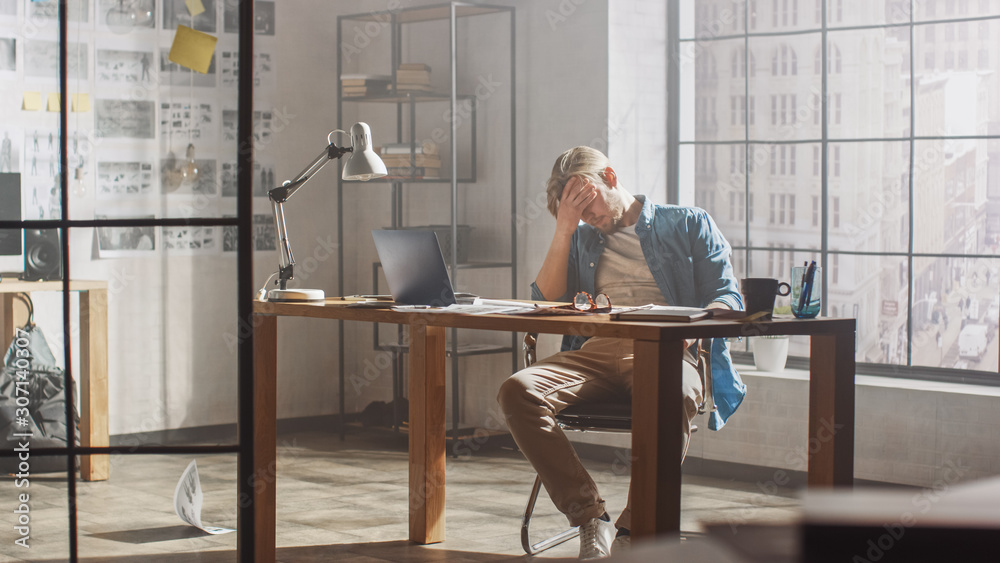 Poster frustrated and tired young creative designer works on laptop at his desk, he browses through concept