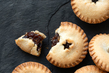 Broken open traditional british christmas mince pie with fruit filling next to whole mince pies on black slate. Top view. Copy space. © Moving Moment