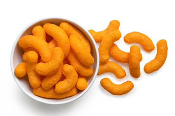 Extruded cheese puffs in a white ceramic bowl next to spilled cheese puffs isolated on white. Top...