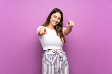 Young woman over isolated purple background points finger at you while smiling