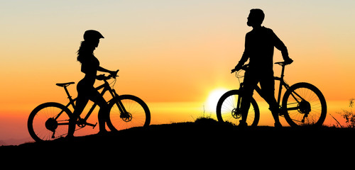 Two silhouettes of cyclists on a sunset background. Athletic guy and a girl on modern mountain bikes go to each other.