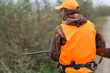 A man with a gun in his hands and an orange vest on a pheasant hunt in a wooded area in cloudy weather. Hunter with dogs in search of game.