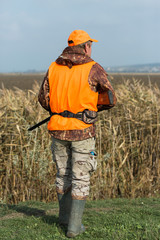 A man with a gun in his hands and an orange vest on a pheasant hunt in a wooded area in cloudy weather. Hunter with dogs in search of game.