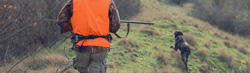 A man with a gun in his hands and an orange vest on a pheasant hunt in a wooded area in cloudy...
