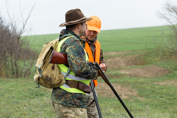 Fototapeta premium A mans with a gun in his hands and an orange vest on a pheasant hunt in a wooded area in cloudy weather. Hunters with dogs in search of game.