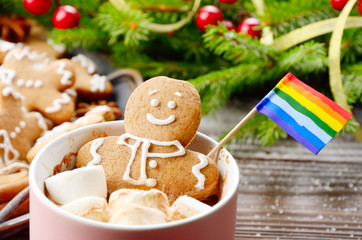 Pink mug with hot chocolate marshmallows and gingerbread man with rainbow flag on background of spruce branch and tray with cookies