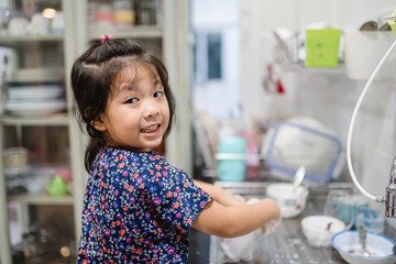 Happy smiley laughing face little cute asian girl washing dishes in kitchen for help her mom.Child...