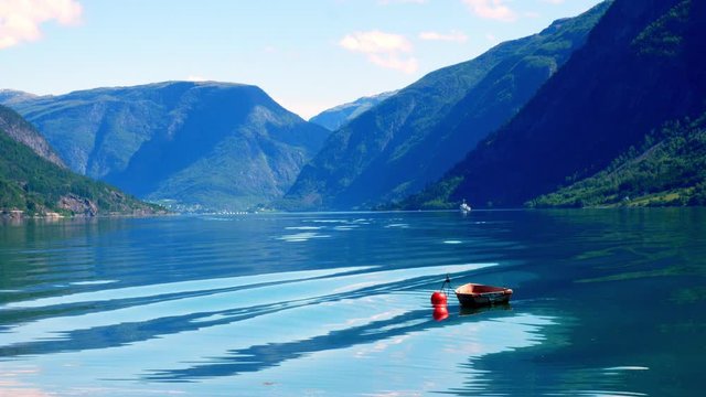 Small wooden boat moored to a red buoy bobs gently in waters of the Lustrafjord in Norway, with reflections of mountains, blue sky and fluffy white clouds. Beautiful Europe Tourism Scenic Footage.
