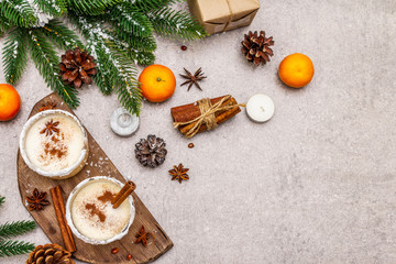 Eggnog with cinnamon and nutmeg for Christmas and winter holidays. Homemade beverage in glasses with spicy rim. Tangerines, candles, gift. Stone concrete background