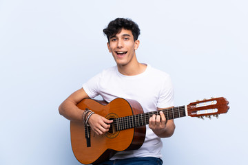 Young Argentinian man with guitar over isolated blue background celebrating a victory