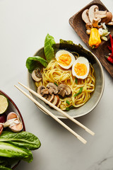 top view of traditional asian ramen in bowl near chopsticks, vegetables on grey surface
