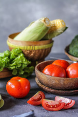 Photo of fresh tomatoes in a bowl on dark background around vegetables, carrot, salt, black pepper, corn, broccoli. Slice tomatoes. Harvesting tomatoes. Drops of water vegetables. Wooden table. Image