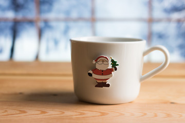 Fototapeta premium Close-up: White Mug and Image of Santa Claus with Christmas Tree on the Wooden Table against Blurry Window with View of Snowy Winter Outside, - Copy Space.