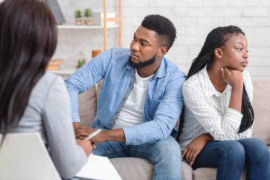 Indifferent black spouses sitting on couch at marital therapist's office.