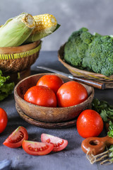 Photo of fresh tomatoes in a bowl on dark background around vegetables, carrot, salt, black pepper, corn, broccoli. Slice tomatoes. Harvesting tomatoes. Drops of water vegetables. Wooden table. Image