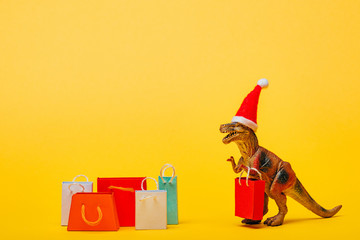 Toy dinosaur in santa hat with shopping bags on yellow background