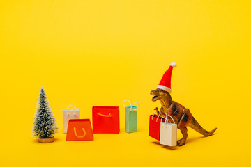 Toy dinosaur in santa hat with shopping bags and christmas tree on yellow background