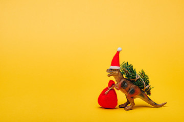 Toy dinosaur in santa hat with sack and fir on yellow background