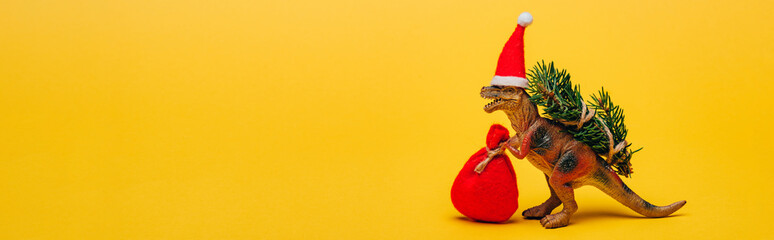 Toy dinosaur in santa hat with sack and fir on yellow background, panoramic shot