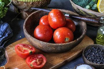 Photo of fresh tomatoes in a bowl on dark background around vegetables, carrot, salt, black pepper, corn, broccoli. Slice tomatoes. Harvesting tomatoes. Drops of water vegetables. Wooden table. Image