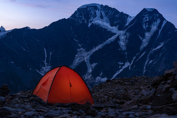 The tent is in the background of the steep cliffs in the snow. Overnight in the mountains. Illuminated tent on the inside.