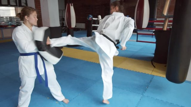 Wide shot of young female kickboxer wearing white kimono kicking punching pillow her female opponent holding in hands