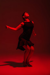 Fototapeta na wymiar sensual dancer in black dress performing tango on dark background with red lighting