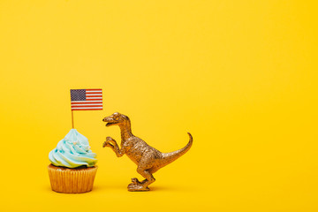 Toy dinosaur beside cupcake with american flag on yellow background