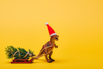 Toy dinosaur in santa hat with fir on sleigh on yellow background