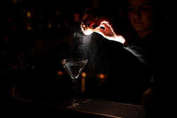 Professional female bartender sprinkling on a alcoholic drink in the cocktail glass with a orange zest juice