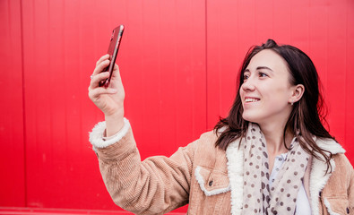 Young woman blogger stands in front of the red wall, holds a mobile phone in her hands and takes pictures, takes a selfie or communicates