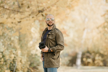Bald photographer with a beard in aviator sunglasses with mirror lenses, olive cargo military jacket, blue shirt with digital wristwatch holds his camera and poses in the forest in the afternoon.