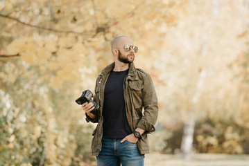 Bald photographer with a beard in aviator sunglasses with mirror lenses, olive military jacket, blue jeans and shirt with digital wristwatch holds the camera by the lens turning his head to the right 