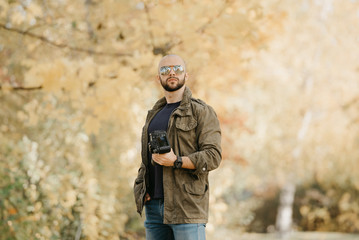 Bald photographer with a beard in aviator sunglasses with mirror lenses, olive military combat jacket, blue jeans and shirt with wristwatch poses holding his dslr camera and looks straight in the fore