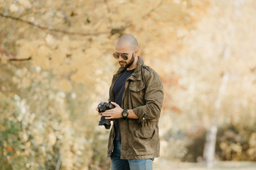 Bald photographer with a beard in aviator sunglasses with mirror lenses, olive cargo military jacket, blue jeans and shirt with digital wristwatch checks system preferences in the camera in the forest