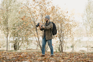 A military photojournalist with a beard in aviator sunglasses with mirror lenses, olive combat jacket, jeans, hat with a backpack with wristwatch holds the DSLR camera, checks photos near the forest - obrazy, fototapety, plakaty