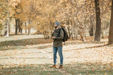 A traveler with a beard in olive military cargo combat jacket, jeans, hat with backpack and wristwatch holds the DSLR camera and the smartphone. A Photographer waits for someone at the noon.