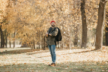 Fototapeta premium A brave photographer with a beard in olive military combat jacket, jeans, red hat with backpack holds the DSLR camera and his wristwatch in the forest at the afternoon