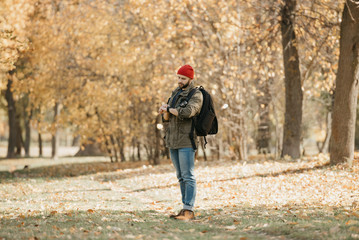 A brave photographer with a beard in olive military combat jacket, jeans, red hat with backpack holds the DSLR camera checks the time on his wristwatch in the forest at the afternoon