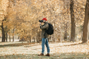 A photographer with a beard in an olive military cargo jacket, jeans, red hat with backpack and wristwatch takes photos with his professional DSLR camera in the forest at the noon