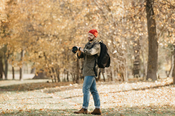 A photojournalist with a beard in an olive military cargo jacket, jeans, red hat with backpack holds his professional DSLR camera and checks the time on the wristwatch in the forest at the noon